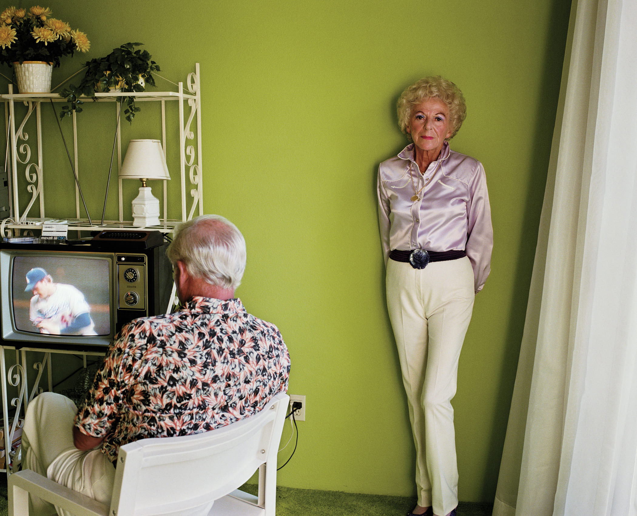 An older man and woman in a room. The man sits watching TV. The woman stands up against the wall looking at the camera.